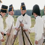 A group of historical reenactors in military uniforms stacking rifles outdoors in a field.
