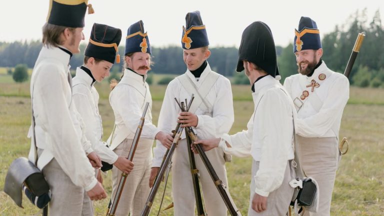 A group of historical reenactors in military uniforms stacking rifles outdoors in a field.